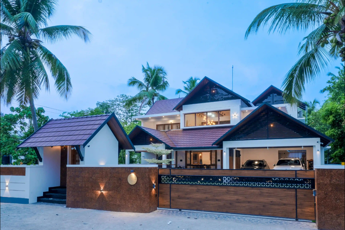 Illuminated home with dark tiled gable roofs, laterite compound wall and timber gate